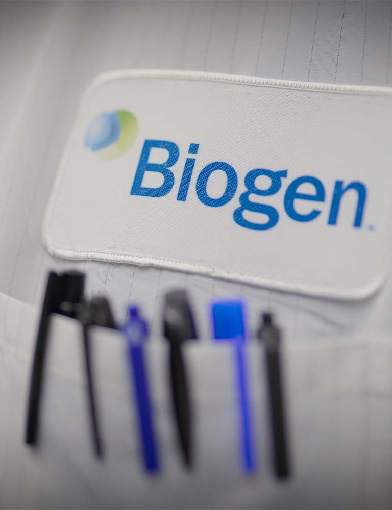Primo piano di un camice bianco con scritta Biogen e numerose penne che spuntano dal taschino